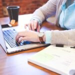 A student typing on a laptop at a wooden desk with a notebook and a cup of coffee