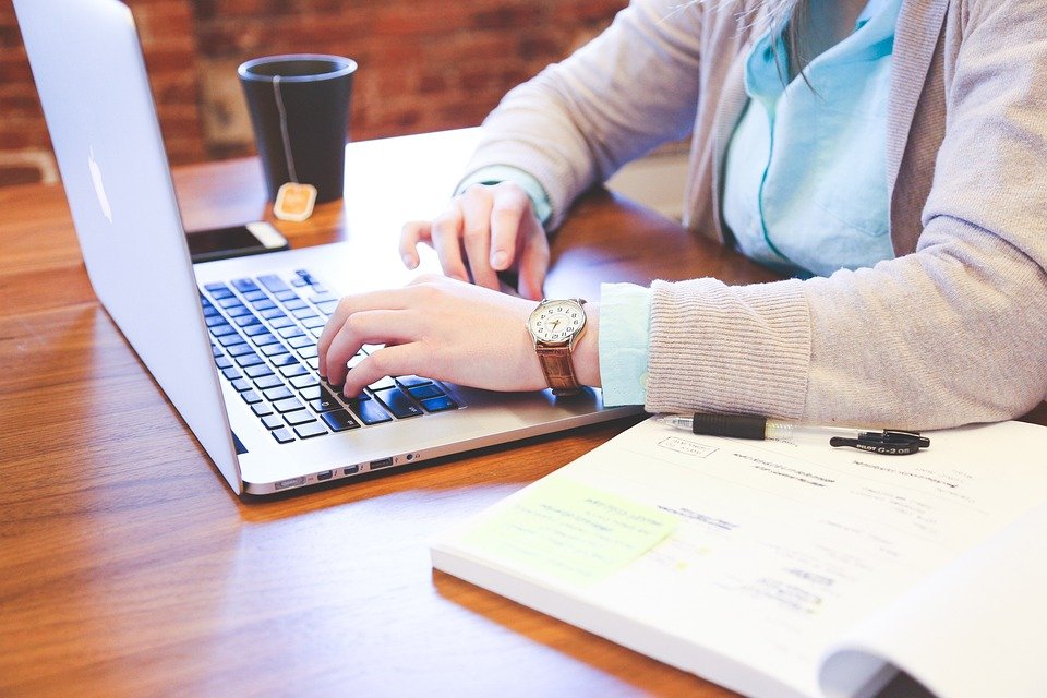 A student typing on a laptop at a wooden desk with a notebook and a cup of coffee